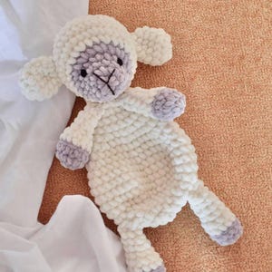 Puede incluir: Un juguete de cordero de peluche hecho a mano en blanco suave y lavanda. El cordero tiene una cabeza redonda con orejas caídas, una cara cosida y un cuerpo texturizado. El juguete está sobre una superficie color melocotón, con una tela blanca en el fondo.