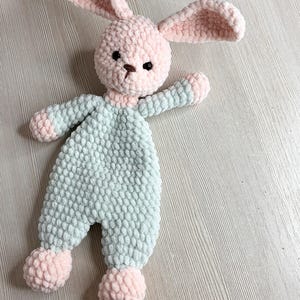 May include: A handmade crochet bunny toy in soft pastel colors. The bunny has a pink head and ears, with a light blue body and pink accents on the feet and hands. The toy has black eyes and a brown nose.