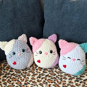 May include: Three handmade crochet cat pillows in pastel shades. Each cat has a different colour scheme, including white, pink, blue, and purple. They feature heart-shaped noses, black whiskers, and large, round eyes. The pillows are on a leopard print surface.