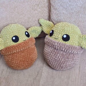 May include: Two plush, handmade, green and brown baby-like dolls. The dolls have large black eyes and small smiles. The dolls are in a pot-like shape, with the head and ears sticking out. The dolls are on a light brown surface.