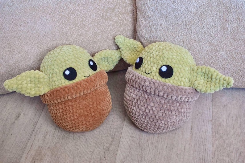 May include: Two plush, handmade, green and brown baby-like dolls. The dolls have large black eyes and small smiles. The dolls are in a pot-like shape, with the head and ears sticking out. The dolls are on a light brown surface.