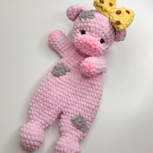 May include: A handmade pink crochet cow-shaped baby blanket with gray spots. The blanket has a yellow bow with brown spots on its head. The blanket is designed to be soft and cuddly.
