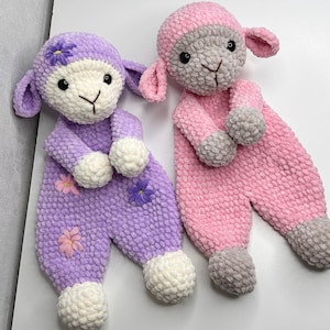 以下が含まれることがあります： 手作りの編みぐるみの子羊のぬいぐるみ2つ。1つは花柄のアクセントが付いたラベンダー色、もう1つはピンク色です。どちらも白い顔、グレーのアクセント、ポンポンの足が付いています。