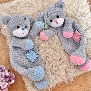 Op de afbeelding: Twee handgemaakte grijze teddyberen met blauwe en roze accenten. De beren hebben gestikte details, waaronder zwarte kruizen en patches op hun lichaam. Ze liggen op een zacht, crèmekleurig oppervlak, wat een gezellige en grillige scène creëert.