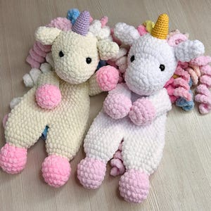 Puede incluir: Dos juguetes de unicornio de peluche. Uno es de color crema, el otro blanco. Ambos tienen detalles rosas, azules y amarillos, incluyendo crines, cuernos y pies de pompón. Los unicornios están tumbados, con las piernas extendidas.