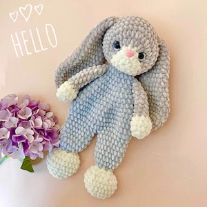 May include: A soft, handmade bunny toy in grey and blue. The bunny has long ears, a white muzzle, and white paws. The word "HELLO" is written in white script above the bunny. A bouquet of purple flowers is in the lower left corner.