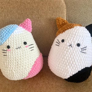 Könnte beinhalten: Zwei handgefertigte, plüschige Katzenkissen. Eine Katze hat blaue und rosa Akzente, die andere braune und schwarze Akzente. Beide haben gestickte Gesichter mit schwarzen Schnurrhaaren und geschlossenen Augen. Die Kissen sind aus weichem, strukturiertem Material.