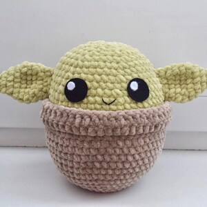May include: A handmade crochet doll resembling a character with large black eyes, large ears, and a smiling face. The doll has a light green head and ears, and a brown body. The doll is sitting on a white surface.