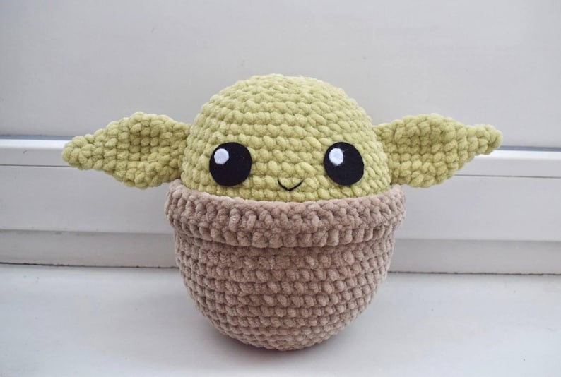 May include: A handmade crochet doll resembling a character with large black eyes, large ears, and a smiling face. The doll has a light green head and ears, and a brown body. The doll is sitting on a white surface.