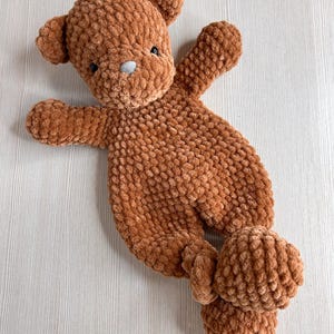 Puede incluir: Un oso de peluche marrón tejido a mano, con un aspecto suave y texturizado. El oso tiene pequeños ojos negros, una nariz gris y brazos y piernas extendidos. El juguete de peluche está diseñado en posición horizontal.