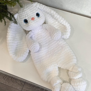 Puede incluir: Un peluche de conejo blanco hecho a mano, con largas orejas, ojos azules y nariz rosa. El conejo está tumbado, con los brazos y las piernas extendidos. El peluche está hecho de hilo suave y texturizado. Mide unos 40 cm de largo.