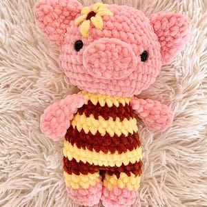 May include: A handmade pink crochet pig stuffed animal with a yellow and brown striped outfit and a yellow flower on its head. The pig has black button eyes and is laying on a fluffy white surface.
