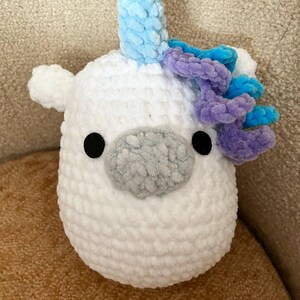 May include: A white, plush unicorn toy with a light blue horn and purple and blue mane. The unicorn has black eyes and a grey nose. The toy is made of soft, textured material.
