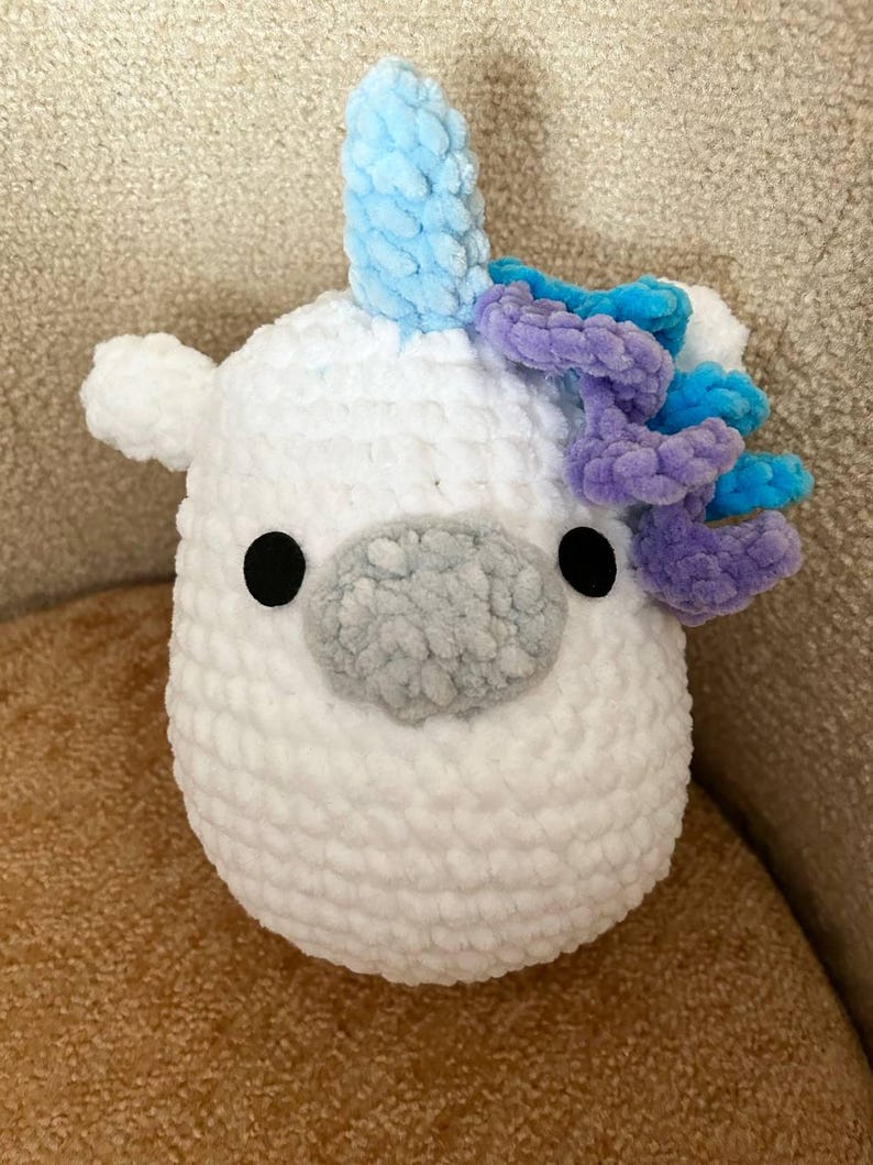 May include: A white, plush unicorn toy with a light blue horn and purple and blue mane. The unicorn has black eyes and a grey nose. The toy is made of soft, textured material.