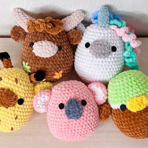 May include: Five colourful, handmade crochet animal plush toys. The collection includes a brown cow, a white unicorn with a rainbow mane, a yellow giraffe, a pink koala, and a green and brown duck. Each has black eyes and is made of soft yarn.