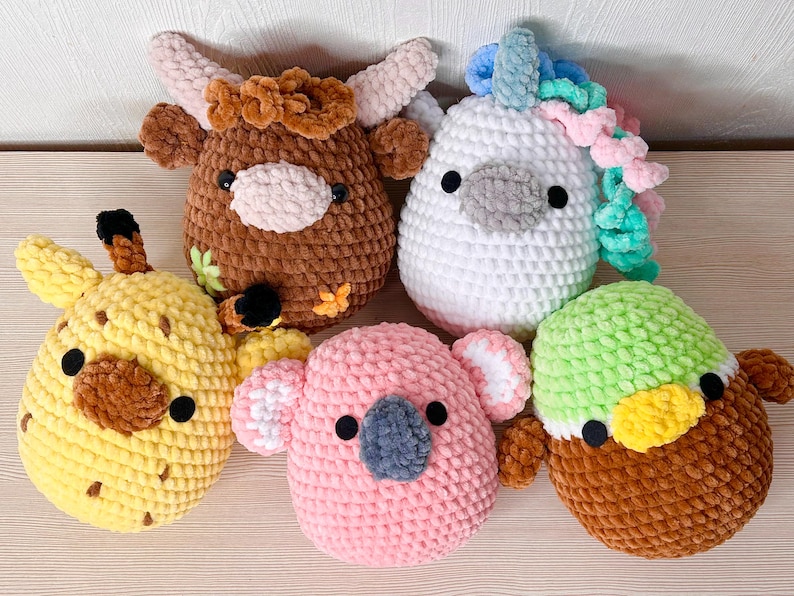 May include: Five colourful, handmade crochet animal plush toys. The collection includes a brown cow, a white unicorn with a rainbow mane, a yellow giraffe, a pink koala, and a green and brown duck. Each has black eyes and is made of soft yarn.