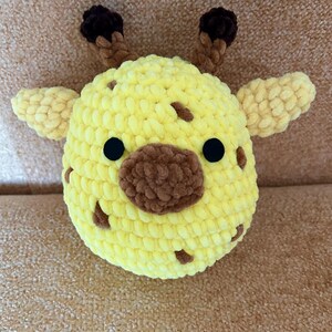 May include: A handmade, plush giraffe head in yellow with brown spots, a brown snout, and black eyes. The giraffe has small, light yellow ears and brown, textured horns. This soft toy is perfect for children.