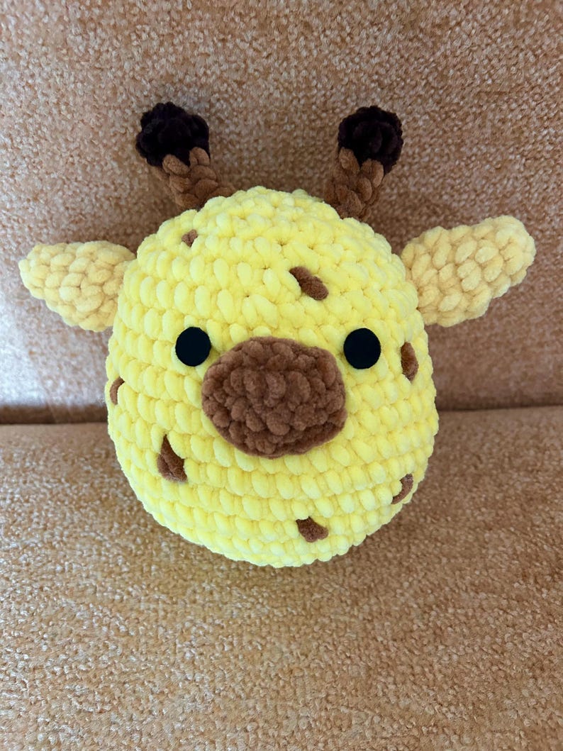 May include: A handmade, plush giraffe head in yellow with brown spots, a brown snout, and black eyes. The giraffe has small, light yellow ears and brown, textured horns. This soft toy is perfect for children.