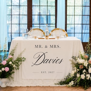 May include: A white wedding table cover featuring the black printed text MR. & MRS. Davies EST. 2027. The table is set with two gold-rimmed chairs, clear glassware, and two large floral arrangements with pink and white roses and green foliage.