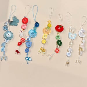 Może przedstawiać: Kolekcja kolorowych, ręcznie robionych zawieszek o różnych wzorach, w tym guziki, motyle i kształty owoców. Każda zawieszka jest nawleczona na biały sznurek, z mieszanką żywych kolorów i zabawnych wzorów.