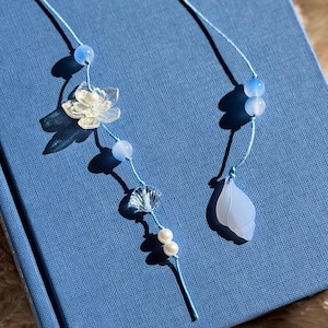May include: Two handmade blue and white beaded bookmarks. One features a clear flower, blue beads, and a shell charm. The other has blue beads and a blue leaf charm. The bookmarks are on a blue background.