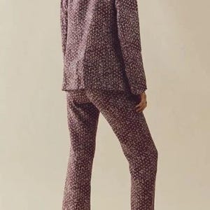 May include: A two-piece floral pajama set in a burgundy color with small white flowers. The set includes a long-sleeved button-up shirt and matching pants. The model is wearing cream-colored loafers. The outfit is made of a soft, comfortable material.