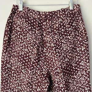 May include: Burgundy-colored pants with a white floral pattern, hanging on a silver hanger. The pants are made of a soft fabric and have a high waist. The floral pattern is small and evenly distributed across the fabric.