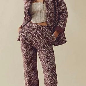 May include: A burgundy floral print suit, including a blazer and matching pants, paired with a cream-colored top and white loafers. The suit features a small, repeating floral pattern. The pants are straight-legged.