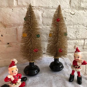 May include: Two gold glitter bottle brush Christmas trees with red, green, and yellow ornaments. Two red and white Santa elf figurines with black boots.