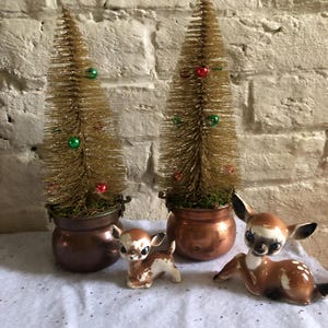 May include: Two gold-colored bottle brush Christmas trees with red and green ornaments sit in copper pots. Two ceramic deer figurines with brown and white markings are in front of the trees. The background is a white brick wall.