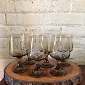 May include: Set of six brown glass wine glasses with a white etched floral design. The glasses are arranged on a wooden slice.