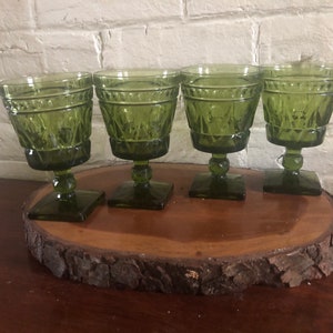 May include: Four green glass goblets with a textured design sit on a wooden slice. The goblets have a square base and a round bowl.