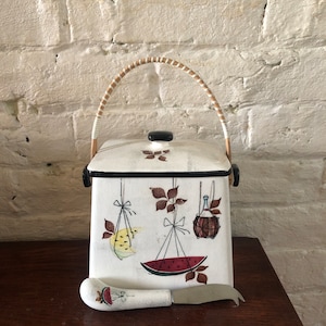 May include: White ceramic cookie jar with a brown wicker handle and a painted design of fruit and a bottle. The jar has a matching white ceramic knife with a red watermelon design on the handle.