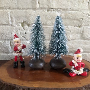 May include: Two small green flocked Christmas trees with brown bases and two ceramic figurines of elves wearing red and white Santa suits on a wooden slice.
