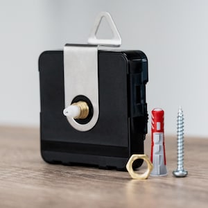 Könnte beinhalten: Ein schwarzes Uhrwerk mit einer silbernen Metallhalterung, einer Messingmutter, einer Schraube und einem rot-grauen Wandanker. Das Uhrwerk hat eine weiße Kunststoffspindel und einen messingfarbenen Ring.
