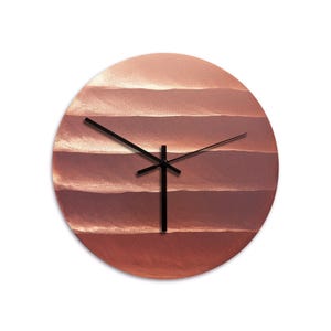 Op de afbeelding: Ronde wandklok met een koperkleurige wijzerplaat met horizontale banden in verschillende tinten koper en bruin. De klok heeft zwarte wijzers en een zwarte centrale paal. Een decoratief item voor thuis of op kantoor, ongeveer 30 cm in diameter.