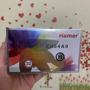 May include: A rectangular box of Hamer candies, held in a hand. The box has a silver border and a colorful design. The text "Hamer" is displayed, along with "E854A9" and "32 candies". The website www.hamer2u.com is also visible.