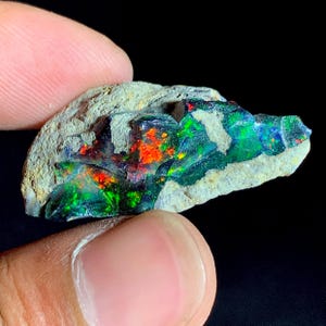 May include: A raw, unpolished opal specimen featuring a dark, textured matrix rock base. The stone displays vibrant flashes of iridescent colour, including intense neon green and bright orange-red patterns embedded within the natural stone surface.