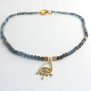 May include: A beaded necklace featuring faceted blue and grey stones, interspersed with small gold-toned metal spacers. The necklace includes a gold-toned toggle clasp and a central gold-toned pendant shaped like an eye with a light blue stone.