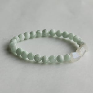 May include: A stretch bracelet composed of smooth, round, pale mint green beads. The design features two faceted, translucent, off-white beads positioned together on one side, creating a subtle contrast in texture and colour.