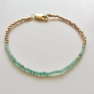 Könnte beinhalten: Ein zartes Armband mit einer Reihe kleiner, facettierter hellgrüner Perlen, kombiniert mit einem Abschnitt aus winzigen, metallisch goldfarbenen Perlen. Das Schmuckstück wird mit einem goldfarbenen Karabinerverschluss geschlossen und bildet einen Kreis.