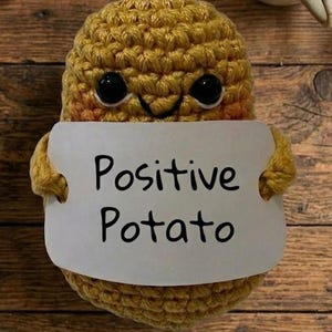 May include: A handmade, crocheted potato toy in shades of yellow and brown, holding a white sign. The sign reads "Positive Potato" in black text. The potato has black button eyes and a stitched smile. The background is a wooden surface.