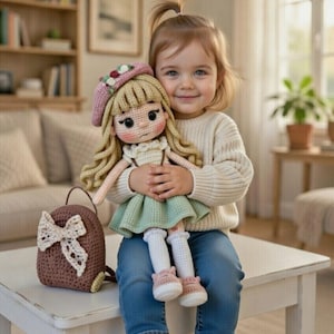 Può includere: Una bambola all'uncinetto fatta a mano con capelli biondi ricci, che indossa un vestito verde e scarpe rosa, tenuta in braccio da un bambino. Uno zaino marrone abbinato con un fiocco color crema è vicino. La bambola ha un basco rosa con accenti floreali.