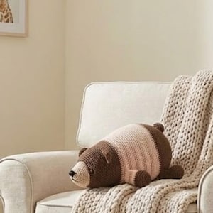 May include: A handmade crochet bear pillow in brown and light pink, lying on a cream-colored chair with a matching knitted throw. The bear has a detailed snout and small ears.