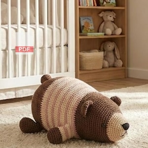May include: A large, handmade crochet bear in shades of brown, cream, and pink lies on a white rug. The bear has a detailed face with a black nose and small ears. The nursery setting includes a crib and a bookshelf with stuffed animals and books.