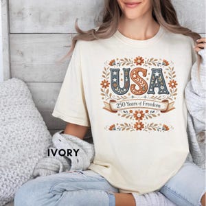 Puede incluir: Camiseta de color marfil con la palabra "USA" en un diseño floral decorativo. El texto "250 Years of Freedom" está en una pancarta debajo de la palabra "USA". La camiseta tiene mangas cortas y cuello redondo.