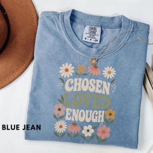Puede incluir: Una camiseta de color azul vaquero con cuello redondo y un estampado gráfico que presenta el texto CHOSEN LOVED ENOUGH rodeado de varias flores coloridas en tonos rosa, amarillo y blanco. La camiseta tiene una etiqueta de la marca Comfort Colors.