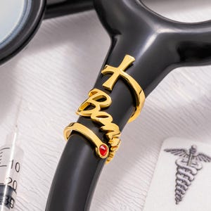 May include: A black stethoscope with gold-colored accents. The accents include a cross, the name "Bon", and a small red gem. A syringe and a medical symbol are visible in the background.