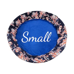 May include: A small, round pet bed with a blue center and a patterned border. The border features a paisley design in shades of orange, red, and white on a dark blue background. The word "Small" is written in white across the center.