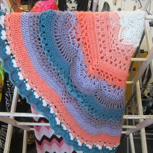 May include: A crocheted blanket with a circular design in shades of teal, purple, pink, and white. The blanket is hanging on a white wooden drying rack.